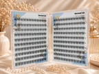 Open clear plastic case displaying two sets of false eyelash clusters labeled 'Mermaid Tail' on light blue backing cards, styled on a draped cream fabric with dried flowers and pearl beads.