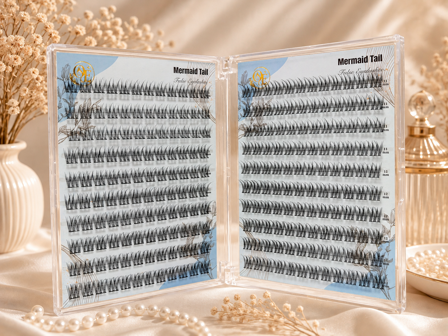 Open clear plastic case displaying two sets of false eyelash clusters labeled 'Mermaid Tail' on light blue backing cards, styled on a draped cream fabric with dried flowers and pearl beads.